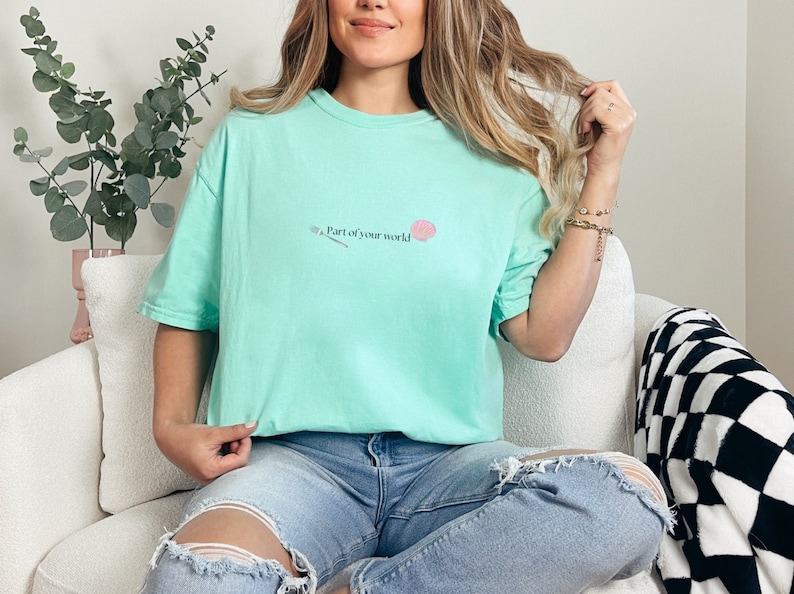 May include: A light blue t-shirt with a pink seashell and a silver fork graphic. The text on the shirt reads "Part of your world."