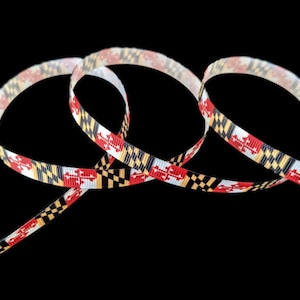 May include: A decorative ribbon with a repeating pattern of red, black, yellow, and white geometric shapes. The ribbon is looped against a black background, showcasing its design. This ribbon is suitable for various crafting and decorative purposes.
