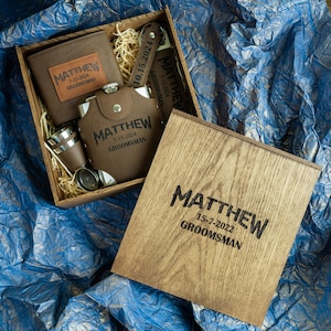 May include: A wooden box with a hinged lid, engraved with the name "MATTHEW" and the date "15-7-2022" along with the word "GROOMSMAN". The box contains a brown leather wallet, a flask, a shot glass, and a keychain.