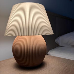 Op de afbeelding: Een moderne tafellamp met een beige geplooide lampenkap en een ronde, geribbelde terracotta voet. De lamp staat op een houten nachtkastje. De lamp is ongeveer 30 cm hoog en geeft zacht omgevingslicht.