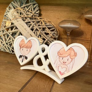 Peut inclure: Un cadre photo blanc en forme de cœur avec un dessin animé de mariés. Le cadre est relié par un symbole esperluette blanc. Le cadre est posé sur une surface en bois. Une décoration en forme de cœur tressé est en arrière-plan.