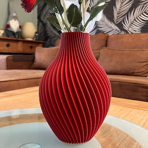 Può includere: Un vaso rosso vibrante con un design unico e scanalato. Il vaso contiene fiori artificiali bianchi e rossi con foglie verdi. Il vaso è su un tavolo di legno con piano in vetro, un elemento di arredo casa moderno.