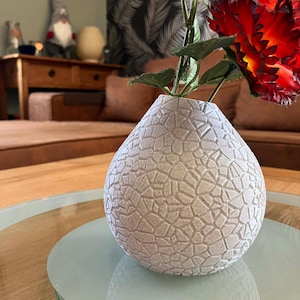 Peut inclure: Un vase sphérique blanc doté d'une surface texturée présentant un motif en mosaïque composé de formes géométriques interconnectées. Le vase possède une ouverture étroite au sommet, d'où émerge une tige unique avec des feuilles vertes et une grande fleur rouge vif.