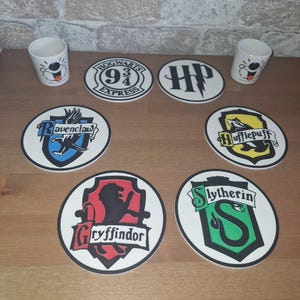 Può includere: Una collezione di sottobicchieri a tema Harry Potter con lo stemma di Hogwarts, il logo HP e gli stemmi delle case di Grifondoro, Serpeverde, Tassorosso e Corvonero. Sono presenti anche due tazze in ceramica bianca con facce di cartoni animati.
