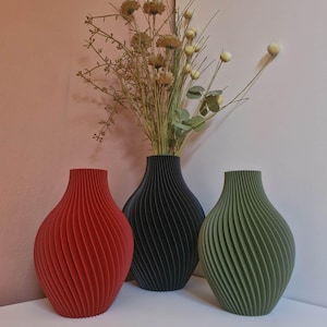 Peut inclure: Trois vases décoratifs rouges, noirs et verts, avec un design côtelé. Les vases sont disposés sur une surface blanche, avec des fleurs séchées dans le vase noir. Les vases ont un design unique et moderne.
