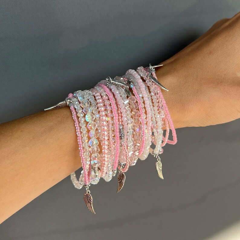 May include: A stack of pink and clear crystal bracelets with silver angel wing charms. The bracelets are layered on a person's wrist.