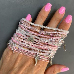 May include: A stack of delicate bracelets featuring pink and clear beads, with silver charms including a hand with the words "Hand Made" and angel wings.