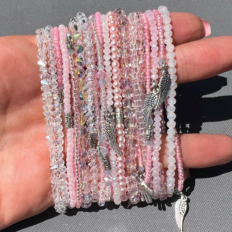 May include: A stack of pink and clear beaded bracelets with silver angel wing charms. The bracelets are layered on top of each other and are tied together with a knot.