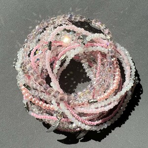 May include: A set of pink and clear beaded bracelets with silver charms. The charms are shaped like wings. The bracelets are stacked on top of each other.