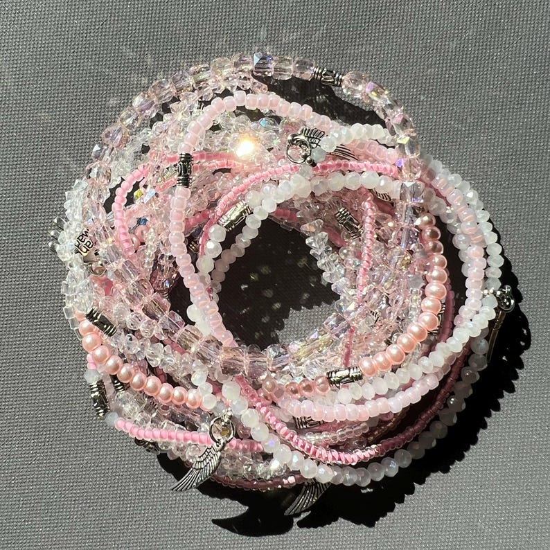 May include: A set of pink and clear beaded bracelets with silver charms. The charms are shaped like wings. The bracelets are stacked on top of each other.