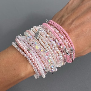 May include: A stack of pink and clear crystal bracelets with a silver angel wing charm. The bracelets are layered on a person's wrist.
