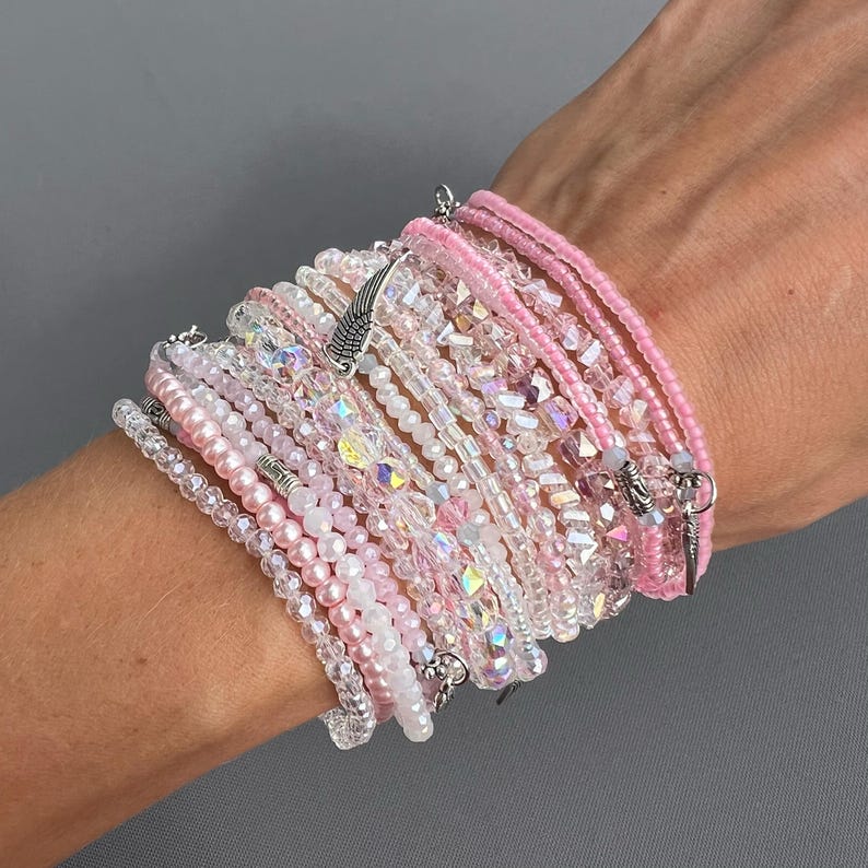 May include: A stack of pink and clear crystal bracelets with a silver angel wing charm. The bracelets are layered on a person's wrist.