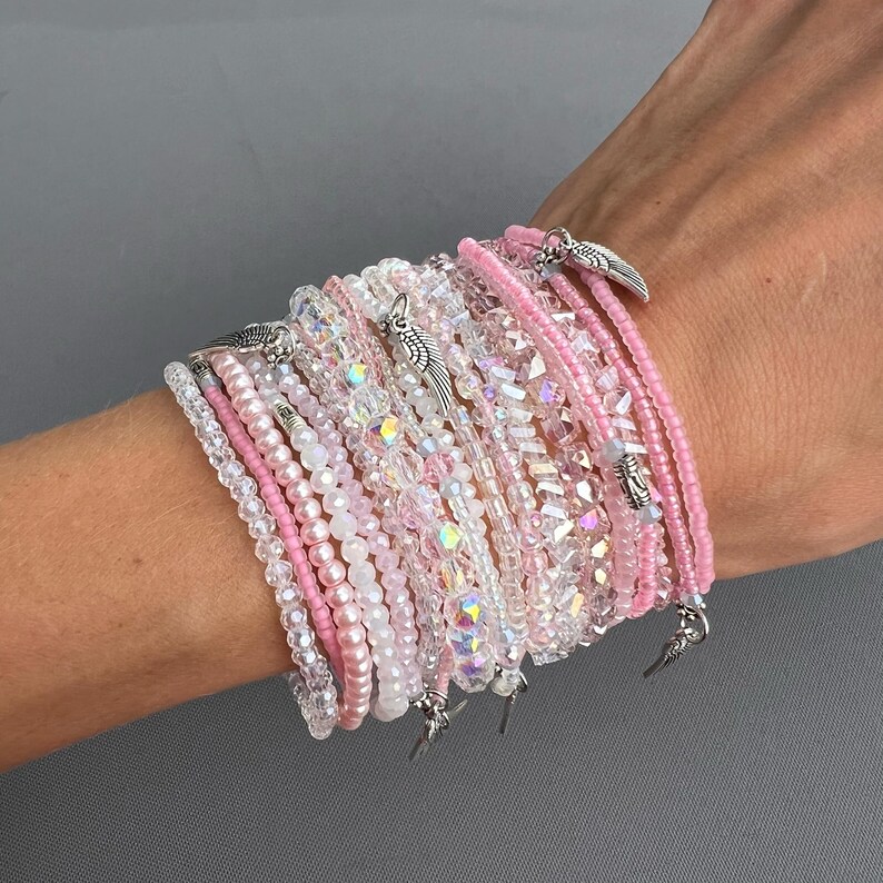 May include: A stack of pink and clear beaded bracelets with silver angel wing charms. The bracelets are layered on a person's wrist.