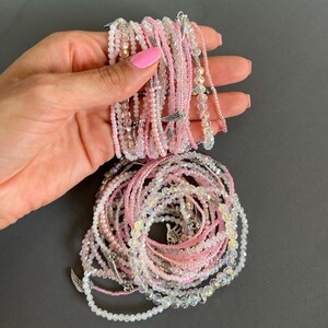 May include: A collection of pink and clear crystal beaded bracelets with silver charms. The bracelets are stacked on top of each other and are held in a hand.