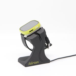 Könnte beinhalten: Ein schwarzer Ladeständer für Smartwatches mit einer limettengrünen und grauen Smartwatch. Der Ständer trägt den Namen "Adrian" in Gold. Das Armband ist schwarz und das Zifferblatt grau.
