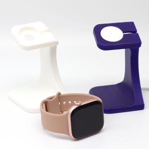 Könnte beinhalten: Eine beige Smartwatch mit schwarzem Bildschirm und roségoldenen Akzenten liegt vor zwei Ladestationen. Ein Ständer ist weiß, der andere tiefviolett. Ein weißes Ladekabel ist sichtbar.