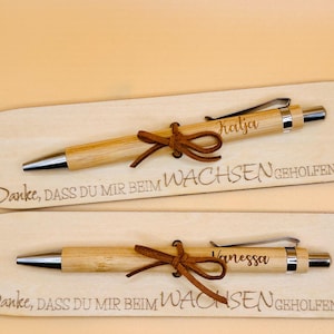 May include: Two personalized bamboo pens with leather bows, each on a wooden plaque. One pen is engraved with "Katja", the other with "Vanessa". The plaques have the text "Danke, DASS DU MIR BEIM WACHSEN GEHOLFEN HAST".