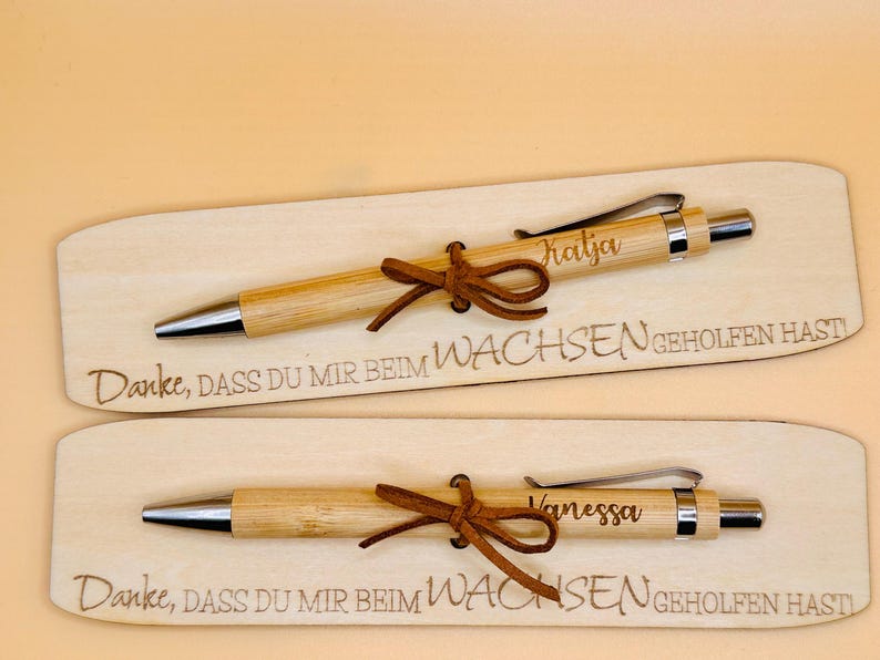 May include: Two personalized bamboo pens with leather bows, each on a wooden plaque. One pen is engraved with "Katja", the other with "Vanessa". The plaques have the text "Danke, DASS DU MIR BEIM WACHSEN GEHOLFEN HAST".