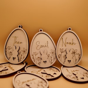 Peut inclure: Ornements d'&oelig;ufs de P&acirc;ques en bois avec des silhouettes de lapins et des noms personnalis&eacute;s, notamment Theo, Emil, Noah, Emma, Lisa et Davi. Les ornements sont de couleur brun clair et ont un aspect rustique et fait main.