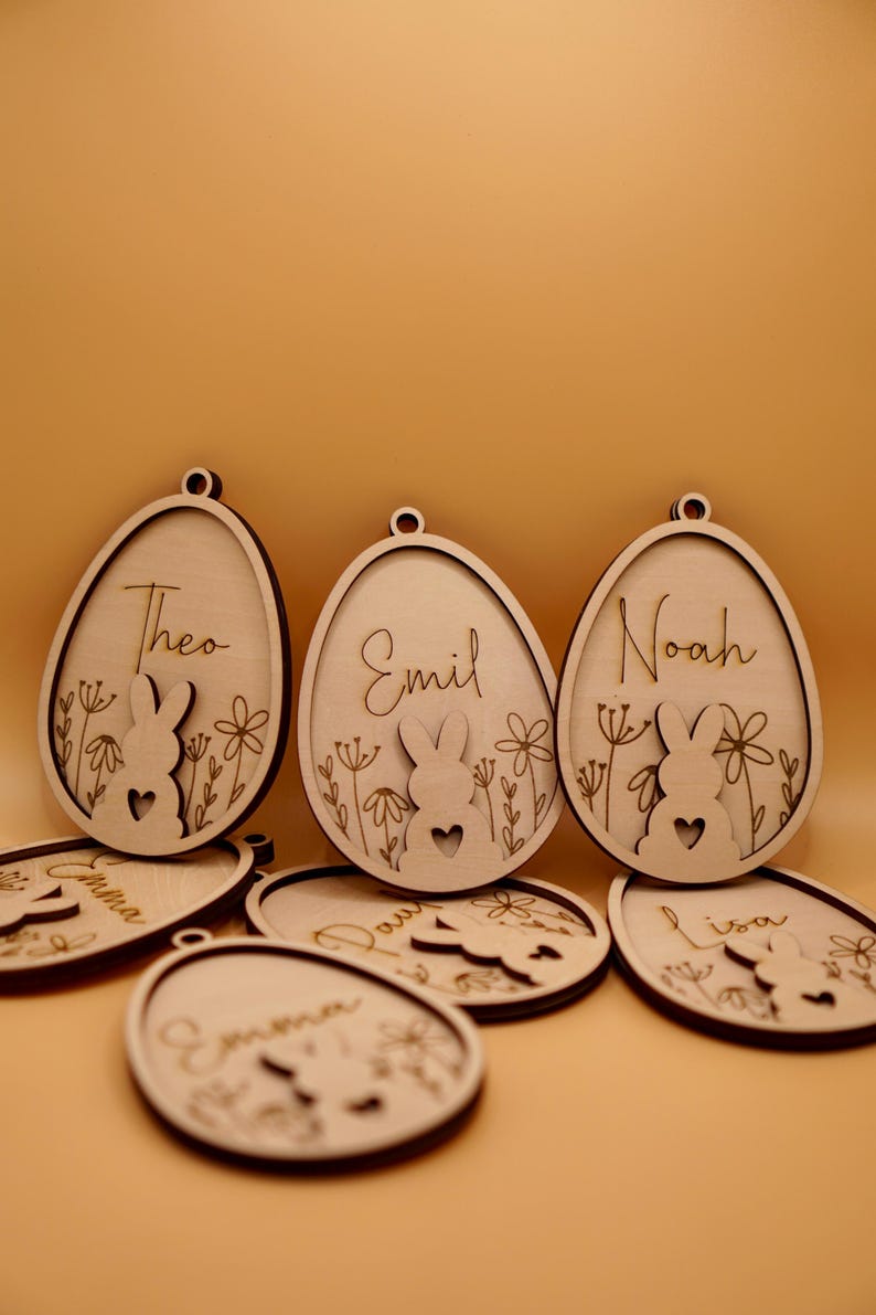 Peut inclure: Ornements d'&oelig;ufs de P&acirc;ques en bois avec des silhouettes de lapins et des noms personnalis&eacute;s, notamment Theo, Emil, Noah, Emma, Lisa et Davi. Les ornements sont de couleur brun clair et ont un aspect rustique et fait main.