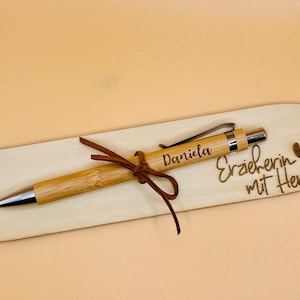 May include: A bamboo pen with silver accents and the name "Daniela" engraved on it, resting on a wooden holder. The holder has the words "Erzieherin mit Herz" and a heart symbol engraved on it. A brown leather cord secures the pen.