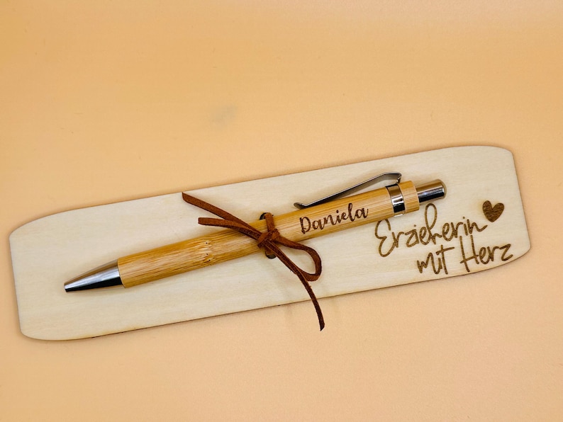 May include: A bamboo pen with silver accents and the name "Daniela" engraved on it, resting on a wooden holder. The holder has the words "Erzieherin mit Herz" and a heart symbol engraved on it. A brown leather cord secures the pen.