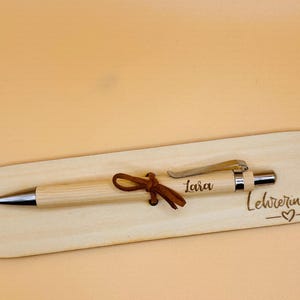May include: A light-colored wooden pen with a silver tip and clip, resting on a matching wooden base. The pen is adorned with a brown leather tie. The base is engraved with the name "Lara" and the phrase "Lehrerin mit".