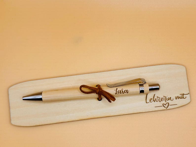 May include: A light-colored wooden pen with a silver tip and clip, resting on a matching wooden base. The pen is adorned with a brown leather tie. The base is engraved with the name "Lara" and the phrase "Lehrerin mit".