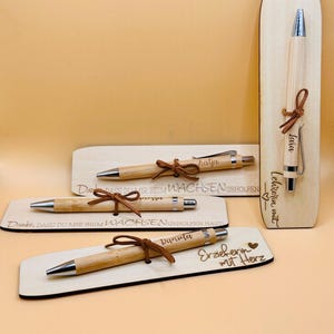 May include: A collection of personalized wooden pens with silver accents and brown leather ties. Each pen is engraved with a name and rests on a wooden base. One base reads "Erzieherin mit Herz."