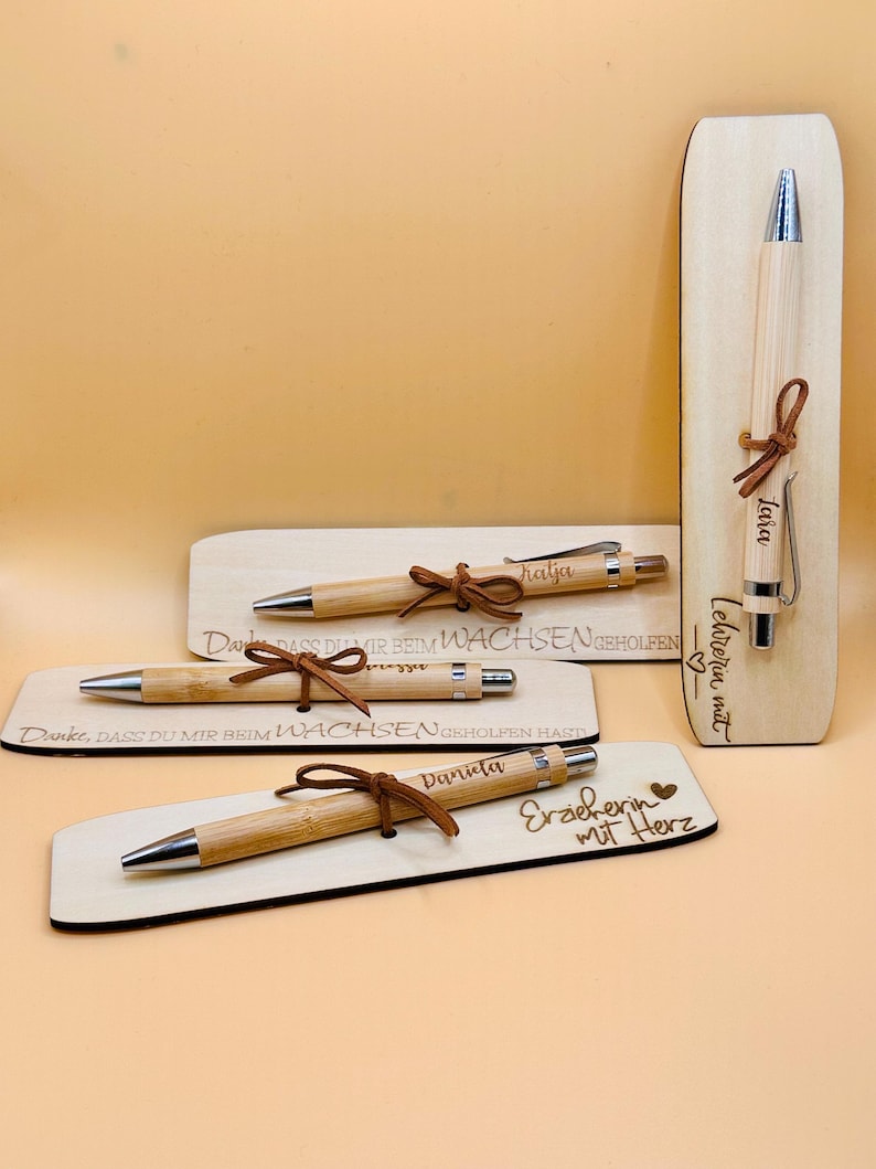 May include: A collection of personalized wooden pens with silver accents and brown leather ties. Each pen is engraved with a name and rests on a wooden base. One base reads "Erzieherin mit Herz."