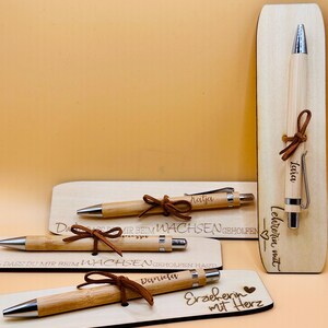 May include: Bamboo pens with silver tips and brown leather accents are displayed with wooden plaques. The plaques have engraved text, including names and phrases like "Teacher with Heart." The pens are tied with leather cords.