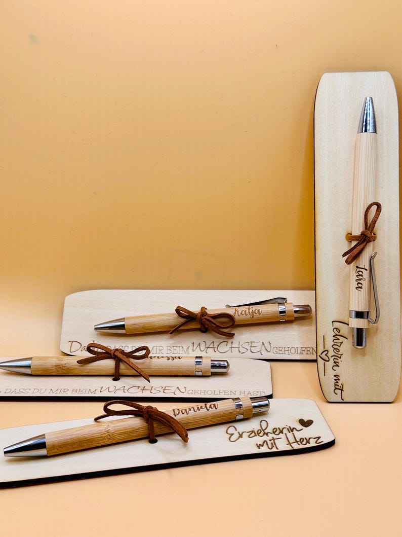May include: Bamboo pens with silver tips and brown leather accents are displayed with wooden plaques. The plaques have engraved text, including names and phrases like "Teacher with Heart." The pens are tied with leather cords.