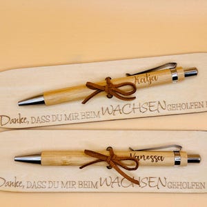 May include: Two wooden pens with silver accents and brown leather ties, each engraved with a name. The pens rest on wooden plaques with the engraved text "Danke, dass du mir beim Wachsen geholfen hast."