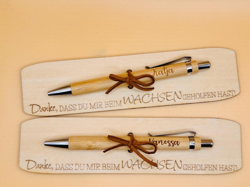 May include: Two wooden pens with silver accents and brown leather ties, each engraved with a name. The pens rest on wooden plaques with the engraved text "Danke, dass du mir beim Wachsen geholfen hast."