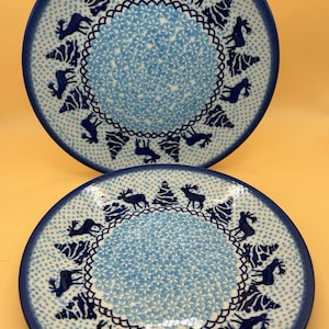 Könnte beinhalten: Zwei Keramikplatten mit blau-weißem Winterdesign. Die Teller haben einen dunkelblauen Rand und ein hellblaues Mittelmuster. Sie sind mit Silhouetten von Elchen und Weihnachtsbäumen verziert.