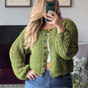 Könnte beinhalten: Eine grüne, geknöpfte Strickjacke mit Lochmuster an den Ärmeln. Der verkürzte Pullover hat einen leuchtenden Grünton und einen gerippten Saum und Bündchen. Die Strickjacke wird mit einer blauen Jeans kombiniert.