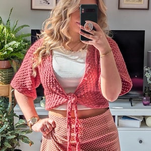 Puede incluir: Un bolero de ganchillo de color coral, de manga corta y cierre de lazo. El suéter tiene un patrón de puntadas texturizado y se usa sobre una camiseta blanca. La persona también lleva un reloj y un anillo dorados.