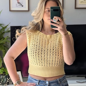 Può includere: Un top senza maniche all'uncinetto giallo chiaro con un design traforato strutturato. Il top presenta un orlo a coste e un piccolo scollo abbottonato. L'indumento è abbinato a jeans blu.