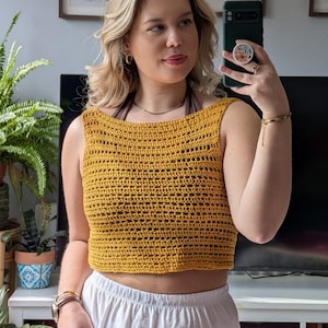 May include: Mustard yellow crochet crop top with a square neckline. The top is sleeveless and has a textured, open weave design. The top is paired with white pants. The image is taken indoors.