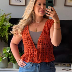 May include: A burnt orange sleeveless crochet vest featuring an open-knit pattern with a V-neckline and a front tie closure. The garment is worn over a solid grey tank top and paired with blue denim jeans, showcasing a textured, lightweight design.