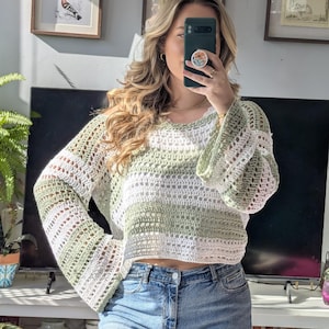 Puede incluir: Un suéter de ganchillo blanco y verde con mangas largas y acampanadas. El suéter presenta rayas horizontales blancas y verde claro. El escote es un simple cuello redondo. El suéter se combina con unos vaqueros azules.