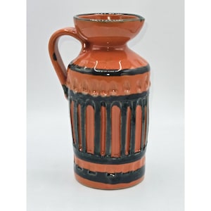 Può includere: Una brocca in ceramica color terracotta con manico e beccuccio largo. La brocca presenta strisce verticali e fasce nere attorno al corpo. Il design è un mix di superfici lisce e strutturate, creando un pezzo visivamente interessante.