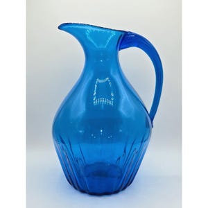 May include: A vibrant blue glass pitcher with a curved handle and a pouring spout. The pitcher has a textured, ridged design on the lower portion of the body. The handle is a solid, curved piece of glass. The pitcher is a decorative item.