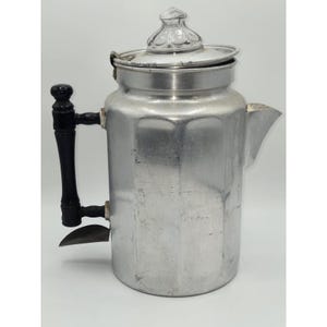 Op de afbeelding: Een vintage aluminium koffiepercolator met een heldere glazen deksel en een zwarte handgreep. De koffiepot heeft een tuit en een metalen filter. De percolator is ontworpen om koffie te zetten.