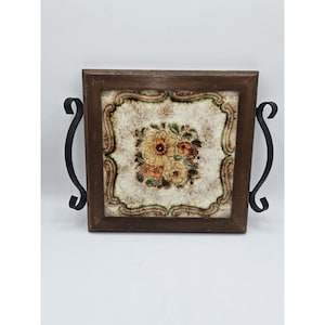 May include: Square wooden trivet with a floral tile insert. The tile features a cream background with a central floral arrangement in shades of orange, yellow, and brown. The trivet has a dark brown wooden frame and black metal handles.