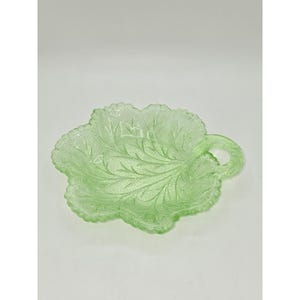 May include: A light green pressed glass dish shaped like a leaf with a textured surface and a small loop handle on one side. The dish features intricate vein details embossed across the interior surface, creating a realistic botanical appearance.