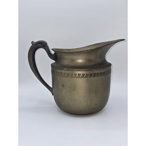 May include: Antique pewter pitcher with a dark handle and a decorative band around the middle. The pitcher has a curved spout and a detailed handle design. The metal has a tarnished appearance, suggesting age and use. The pitcher is likely used for serving beverages.