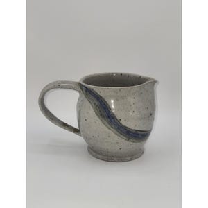 May include: A ceramic creamer pitcher with a speckled light gray finish and a single curved handle. The pitcher features a prominent, hand-painted diagonal blue and gray brushstroke accent across the body and a small spout for pouring liquids.