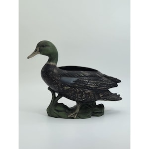 May include: A ceramic duck planter with a dark green head and a black body. The duck is perched on a green base, with a hollowed-out area on its back for planting. The planter has a detailed, textured surface.