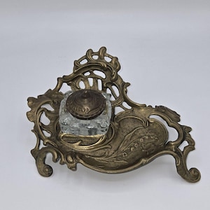 May include: An ornate brass inkwell holder with a glass inkwell. The holder features a floral design with a lily of the valley motif.